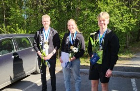 Kolme hymyilevää nuorta opiskelijaa seisomassa vierekkäin parkkipaikalla.