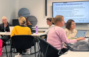 Henkilöt istuvat pöytien ääressä ja keskustelevat
