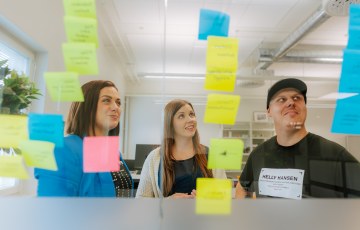 Kolme ihmistä toimistohuoneessa ison ikkunan edessä. Ikkunassa on kiinni postit-lappuja. 