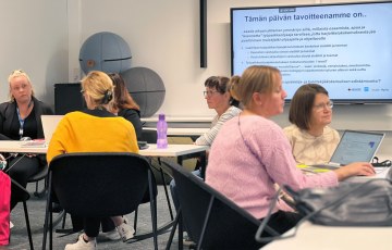 Henkilöt istuvat pöytien ääressä ja keskustelevat