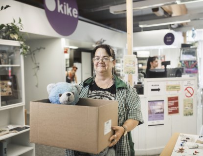 Henkilö seisoo kike-myymälässä ja pitelee käsissään laatikkoa, josta kurkkaa nalle