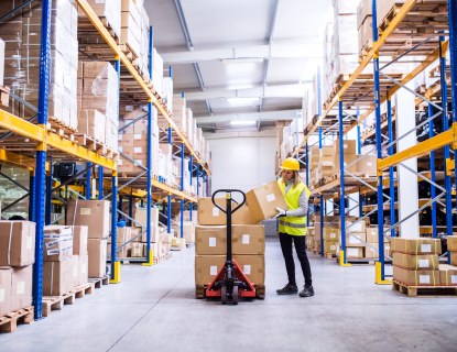 Kuva logistiikan varastosta, jossa naistyöntekijä laittaa isoja paketteja kärrylle.